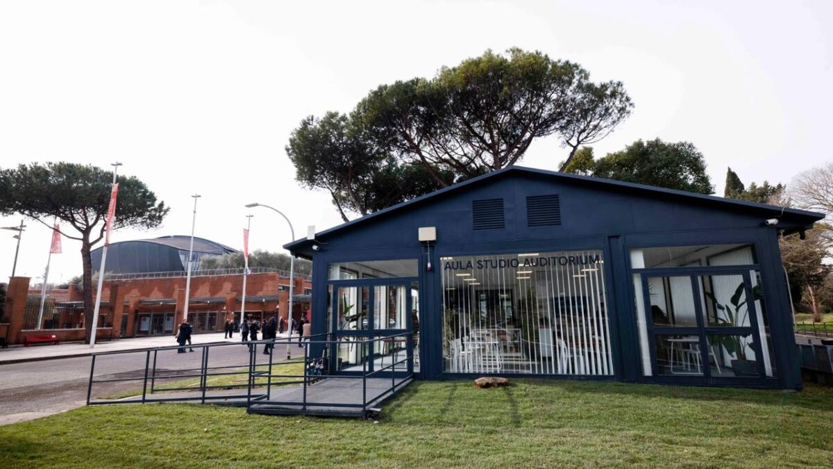 Facciata esterna completa dell'Aula Studio Auditorium: edificio in acciaio blu scuro con pareti vetrate e insegna. Rampa di accesso sul lato sinistro. Sullo sfondo, il Parco della Musica Ennio Morricone con persone in transito.