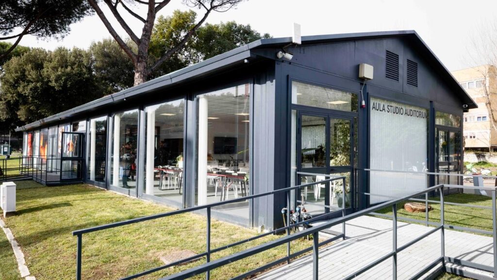 Esterno dell'Aula Studio Auditorium di giorno: struttura in acciaio antracite con pareti in vetro e insegna con il nome. Rampa di accesso per persone con disabilità motoria sul lato destro.