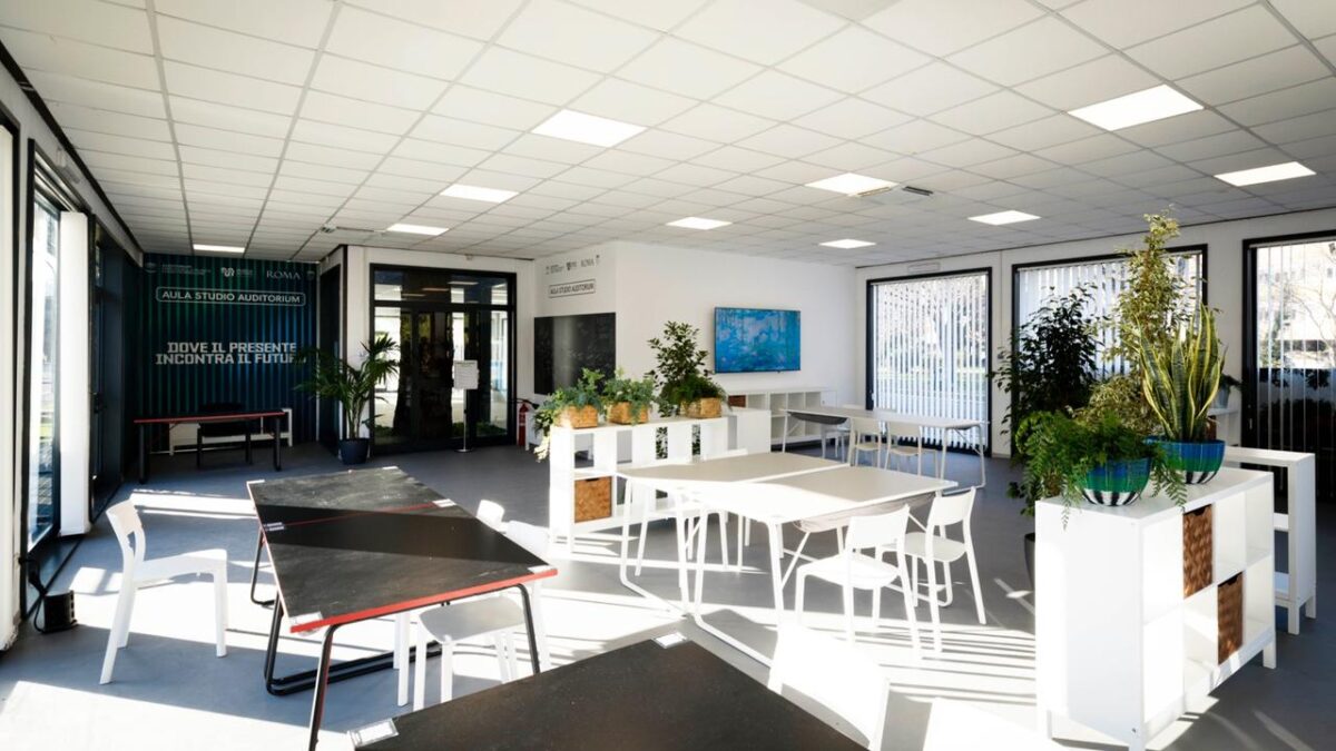 Interior of the Auditorium Study Room: black and white tables, white chairs, open shelving, plants, and a wall-mounted TV screen. Glass entrance doors in the background.