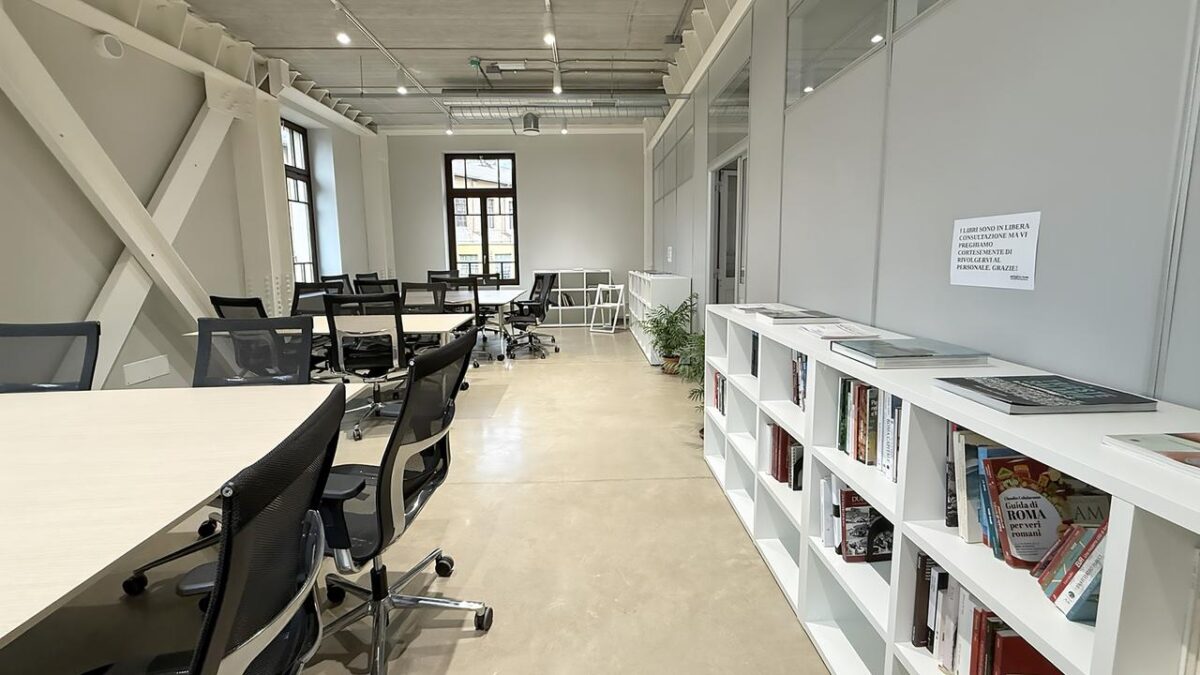 Interior of the Urban Center Metropolitano: long white tables with black ergonomic chairs, white bookshelves with books along the right wall. Industrial ceiling with track lighting.