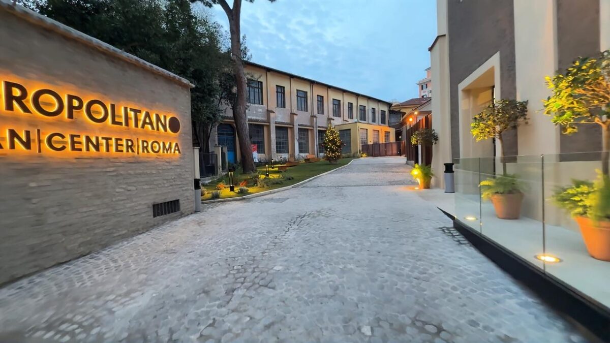 Cortile interno del Metropolitano Urban Center Roma al tramonto. Vialetto in sampietrini illuminato da faretti a terra. Sulla sinistra, insegna luminosa con la scritta Metropolitano Urban Center Roma. Sullo sfondo, edificio su due piani.