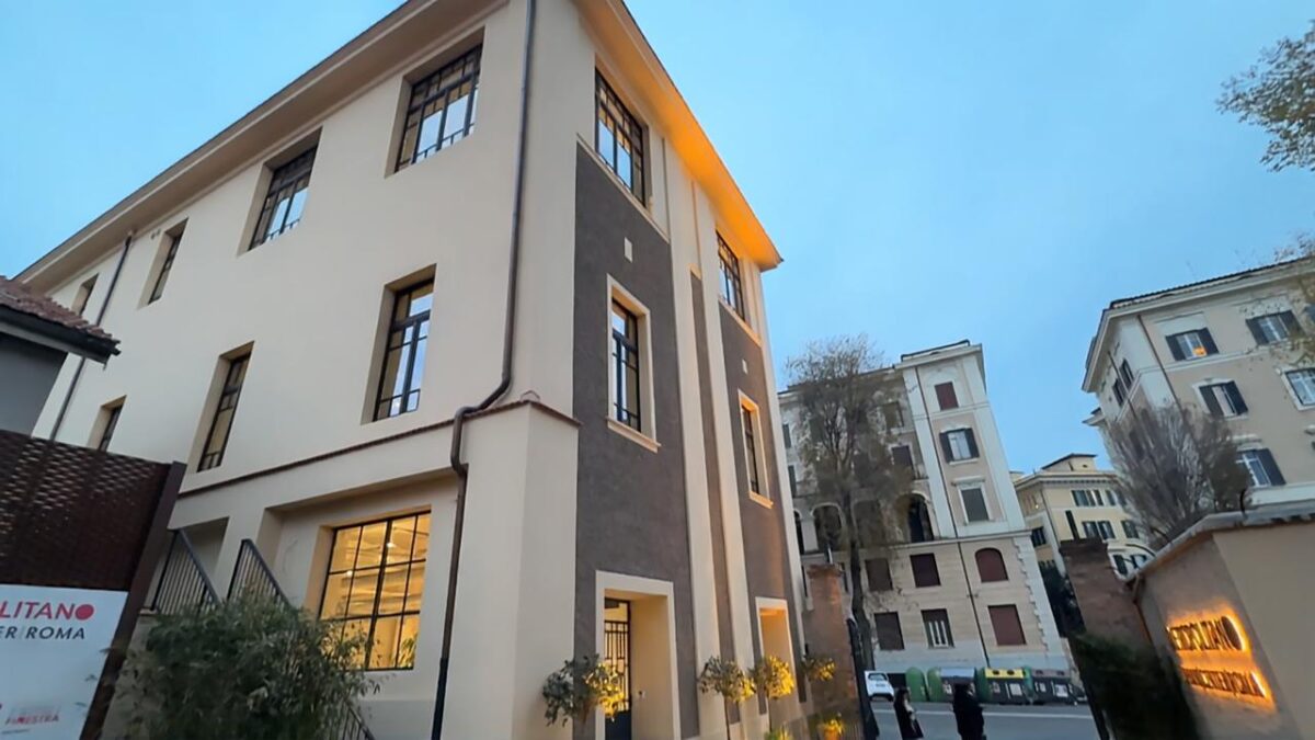 Facciata esterna dell'Urban Center Metropolitano di sera. Edificio su tre piani con intonaco bianco e fasce grigio scuro, finestre con infissi neri. Interni illuminati visibili al piano terra. Insegna Metropolitano Urban Center Roma sul lato sinistro.