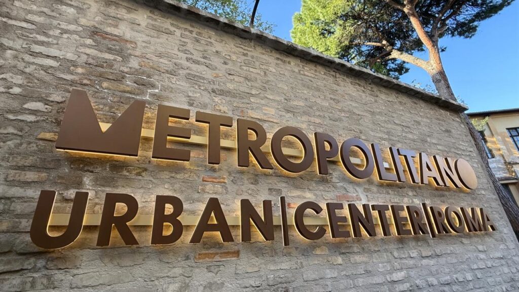 Insegna in lettere tridimensionali color bronzo su muro in mattoni a vista. Testo: Metropolitano – Urban Center Roma. Sullo sfondo, cielo azzurro e cima di un pino romano.