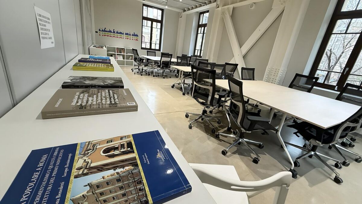 Vista panoramica della sala studio: tavoli bianchi con volumi di architettura e urbanistica in primo piano, file di sedie ergonomiche nere sullo sfondo. Soffitto alto con travi bianche a vista e ampie finestre. Logo Metropolitano sulla parete di fondo.