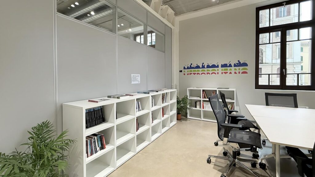 Area lettura con scaffalature bianche a muro contenenti libri. Sulla parete logo illustrato e scritta Metropolitano in stile grafico colorato. Finestra con vista su edifici esterni. Tavolo da studio con sedie ergonomiche nere.